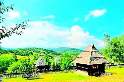 Традиционална златиборска кућа брвнара са високим стрмим кровом од шиндре, смештена на пропланку окружена касним поподневним сунцем